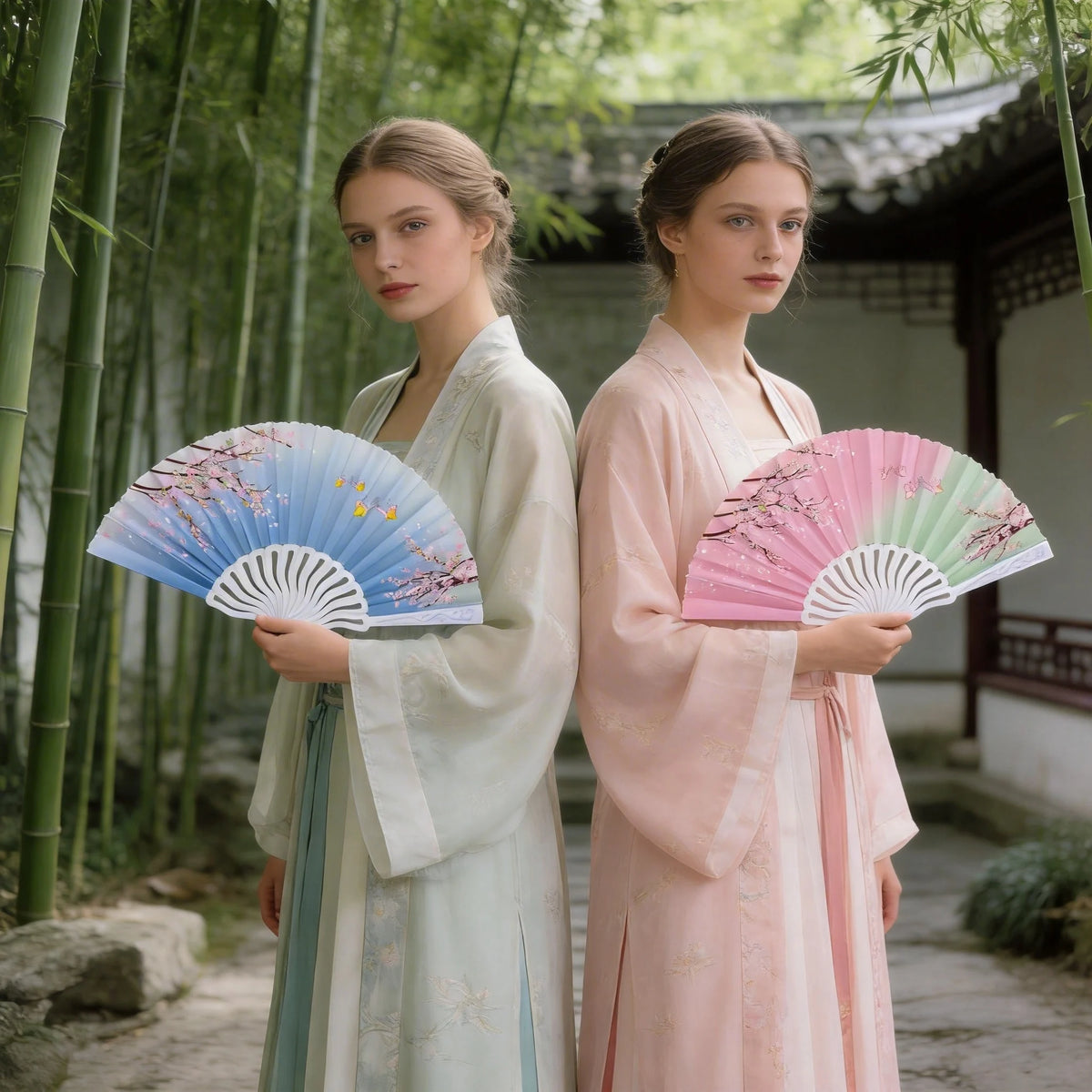 Two women in traditional clothing hold pink and blue printed plastic folding fans in a lush garden setting.