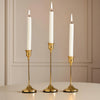 A set of three elegant gold metal candlesticks with white candles displayed on a light surface.