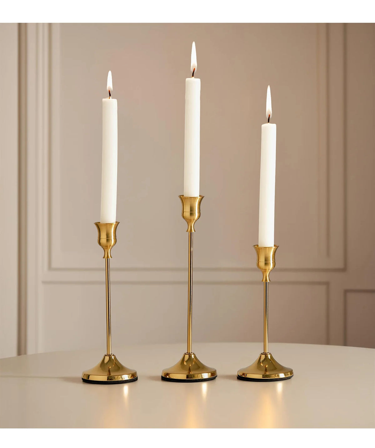 A set of three elegant gold metal candlesticks with white candles displayed on a light surface.