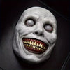 A realistic smiling demon latex mask with pale gray skin, glowing eyes, and a wide, toothy grin.