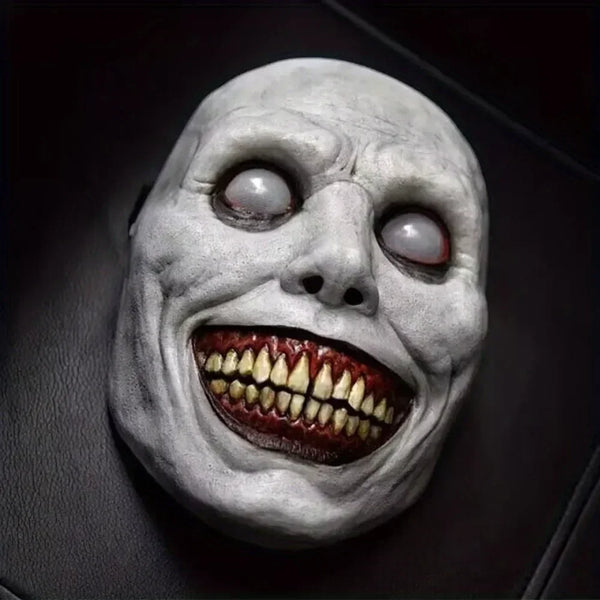 A realistic smiling demon latex mask with pale gray skin, glowing eyes, and a wide, toothy grin.