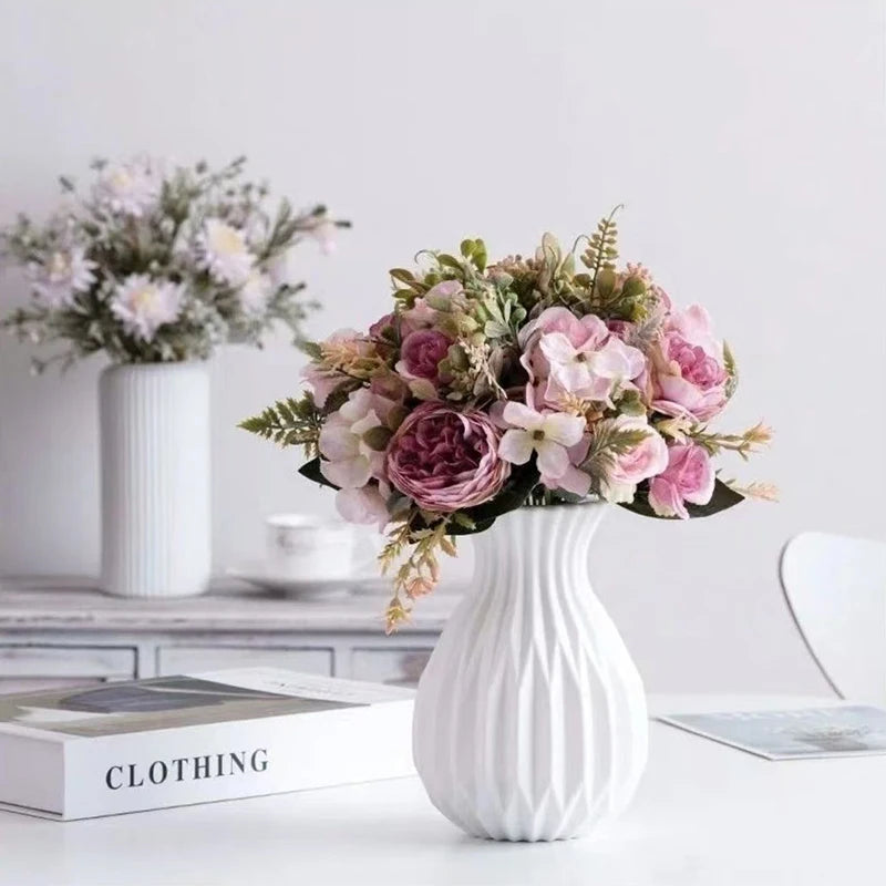 A white ceramic flower vase filled with pink and purple floral arrangements on a white surface.