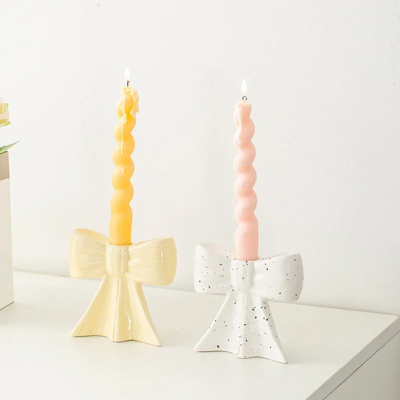 A pair of elegant ceramic bow candle holders, one yellow and one pink, displayed on a white surface.