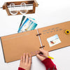 A hand is writing in a brown kraft scrapbook album with photos and a wooden frame nearby.