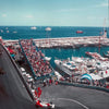 Aerial view of a motorsport racing track with a large crowd and boats docked in the harbor under a clear blue sky.