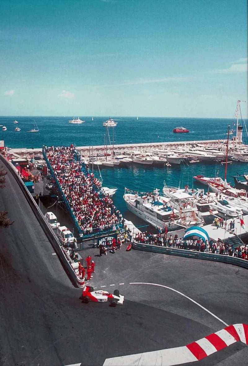 Aerial view of a motorsport racing track with a large crowd and boats docked in the harbor under a clear blue sky.