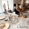 A display of three metal taper candle holders with lit candles on a white tablecloth.