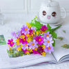 A vibrant bouquet of purple, pink, and yellow artificial daisy flowers displayed on an open book next to a cute ceramic teapo