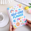 A yellow spiral notebook titled Self-Care Planner with colorful stickers is held by a hand on a white surface.