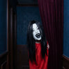 A person wearing a white latex horror mask with long black hair and a red shirt stands in a dimly lit room.