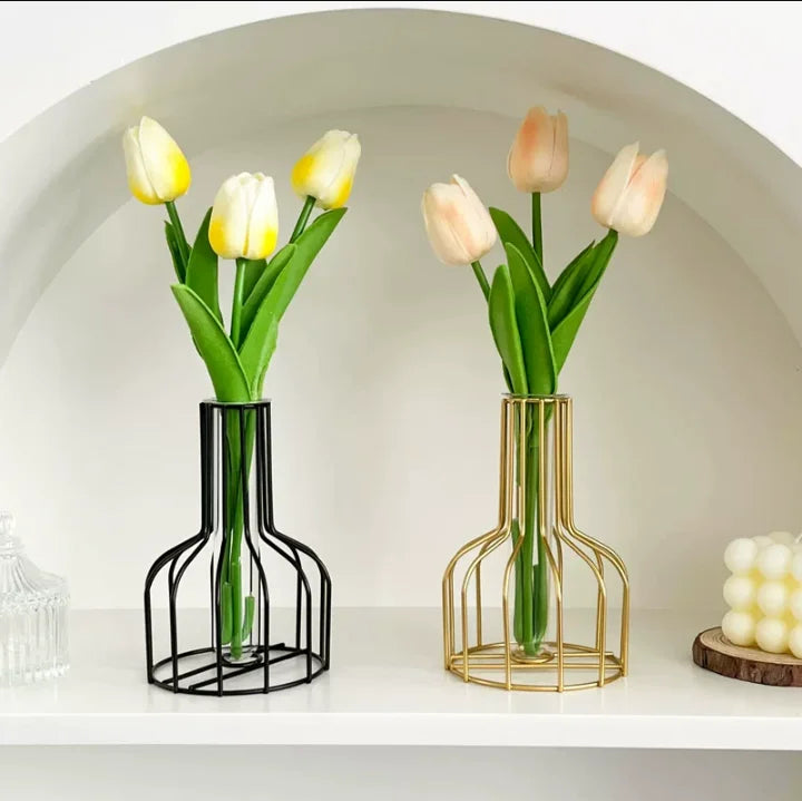 A pair of handcrafted iron hollow vases holding pale yellow and peach tulips on a white shelf.