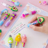 A hand holds a set of five cute cartoon mini ballpoint pens in various colors and designs on a white grid paper.
