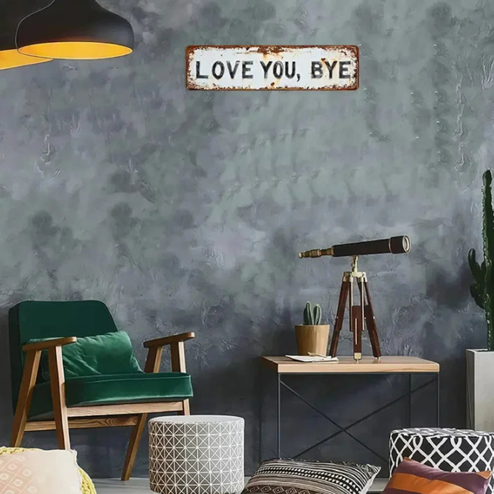 A rustic metal sign reading LOVE YOU BYE mounted on a gray textured wall above a wooden chair and plant.