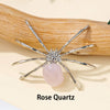 A handcrafted crystal spider figurine with silver legs and a pink rose quartz body resting on a wooden surface.