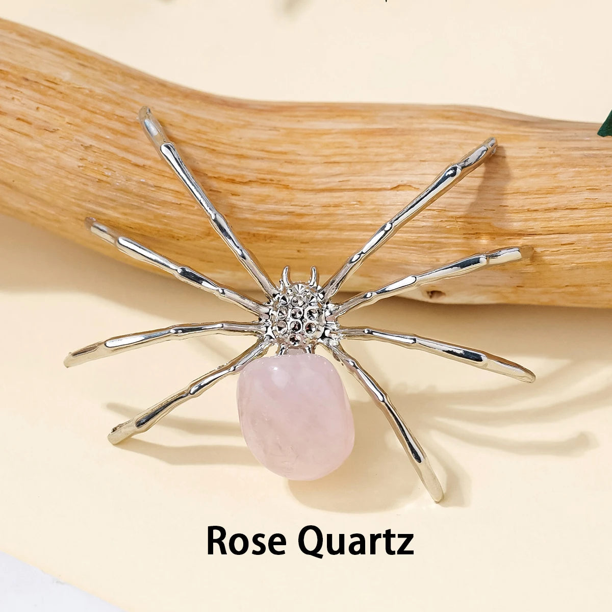 A handcrafted crystal spider figurine with silver legs and a pink rose quartz body resting on a wooden surface.