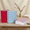 A stack of pink and white photo albums rests on a wooden table next to a vase with dried foliage.