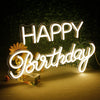 A glowing neon sign spelling out HAPPY BIRTHDAY is displayed on a surface with green foliage in the background.