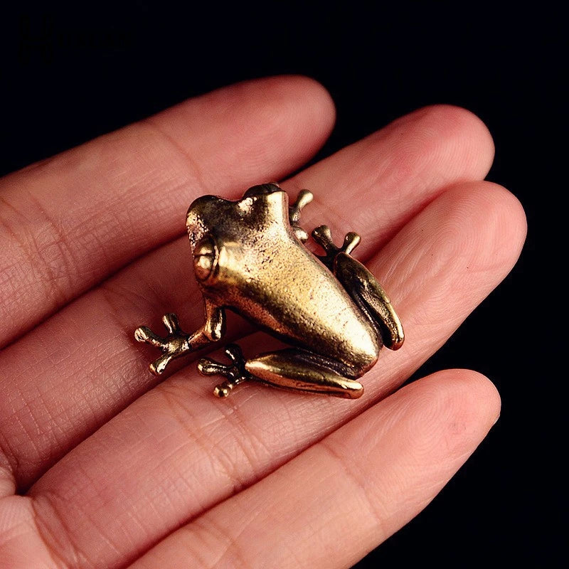 A vintage brass frog statue ornament resting on a human hand against a dark background.