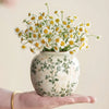 A hand holds a white ceramic vase filled with yellow and white daisies.