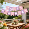 A pink and purple birthday garland banner is strung across a porch, decorated with flowers and greenery.