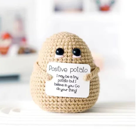 A handmade knitted plush potato doll with a positive message tag sitting on a white surface.