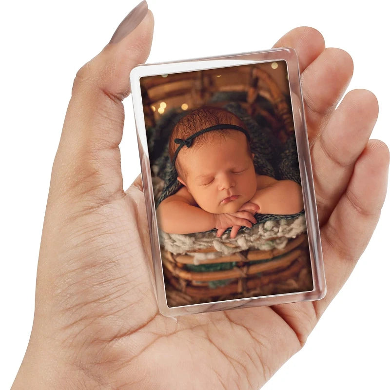 A hand holds a magnetic mini photo frame displaying a sleeping newborn baby in a woven basket.