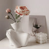 A white ceramic vase shaped like an artistic human form holds a pink rose flower.