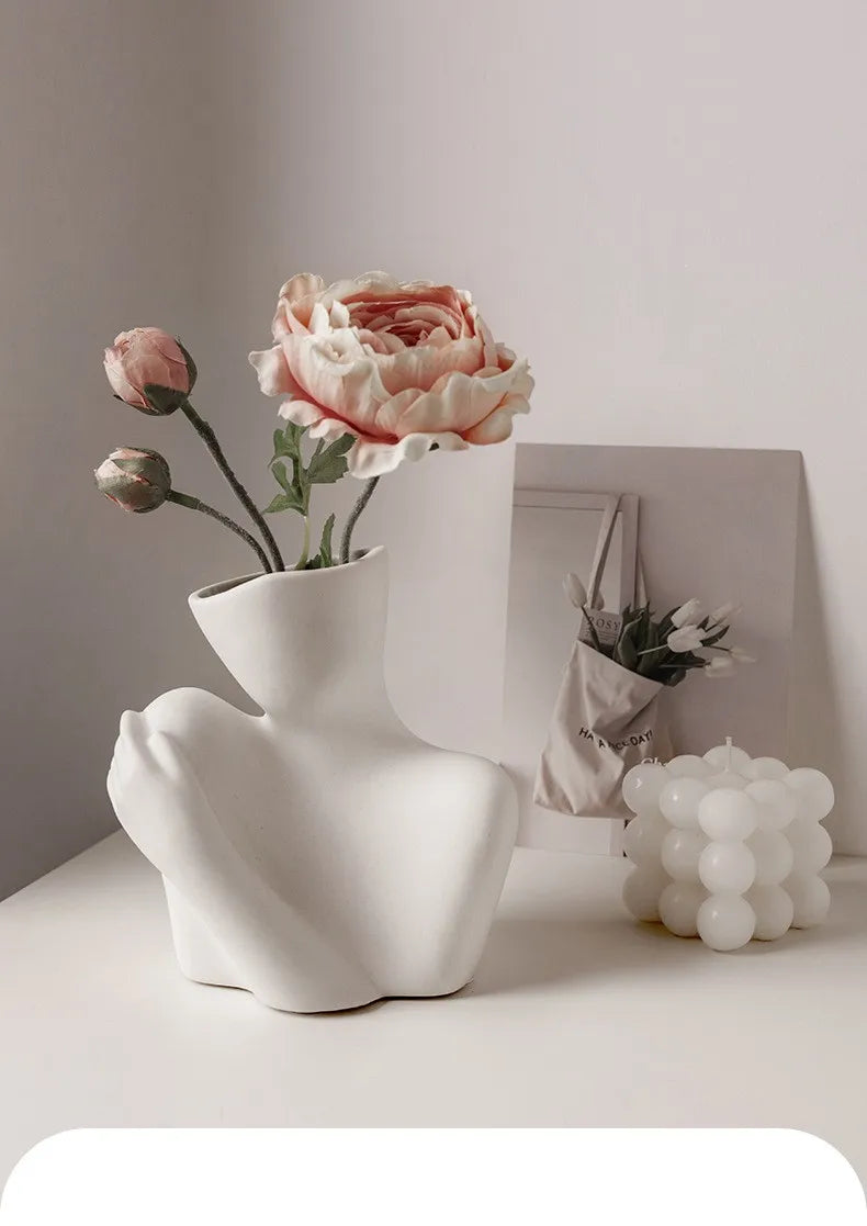 A white ceramic vase shaped like an artistic human form holds a pink rose flower.