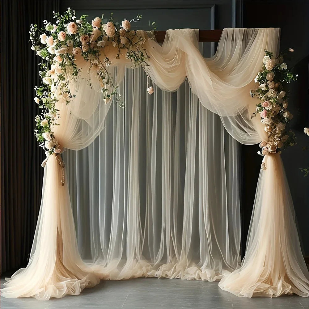 A sheer beige tulle fabric backdrop with draped curtains and floral accents for wedding or event decor.