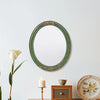 An oval wall mirror with a green frame and intricate lace design hangs on a white wall next to a white rose.