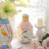 A pink and white striped ceramic vase with floral designs sits next to a yellow candle and soft white fabric.