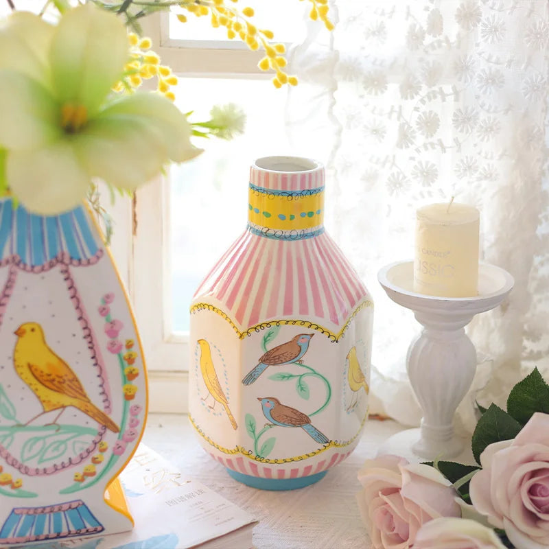 A pink and white striped ceramic vase with floral designs sits next to a yellow candle and soft white fabric.