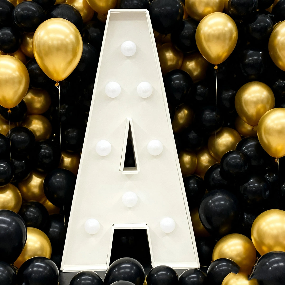 A large white marquee letter shaped like the letter A is displayed amidst a dense background of gold and black balloons.
