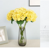 A bouquet of bright yellow artificial hydrangea flowers in a clear glass vase on a light surface.