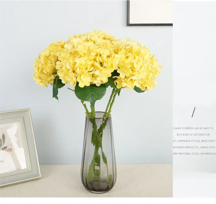A bouquet of bright yellow artificial hydrangea flowers in a clear glass vase on a light surface.