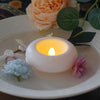 A white LED floating sensor tealight candle is lit on a plate surrounded by green leaves and light pink flowers.