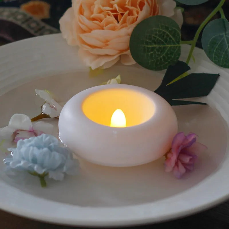 A white LED floating sensor tealight candle is lit on a plate surrounded by green leaves and light pink flowers.