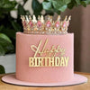 A pink cake with a gold acrylic happy birthday topper featuring a crown and text.
