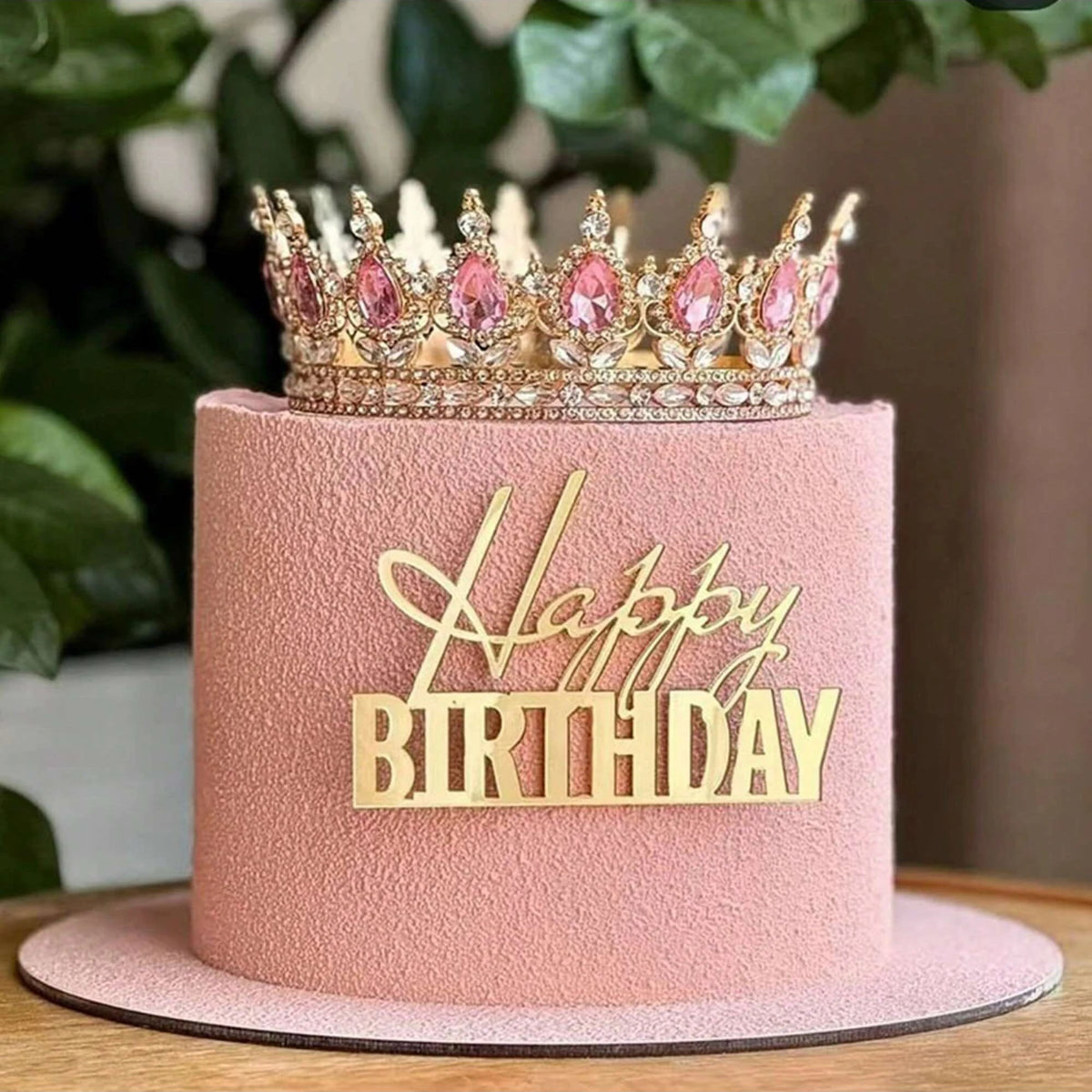 A pink cake with a gold acrylic happy birthday topper featuring a crown and text.