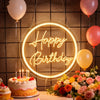 A glowing neon sign spelling Happy Birthday is displayed on a dark wall with balloons and a cake.