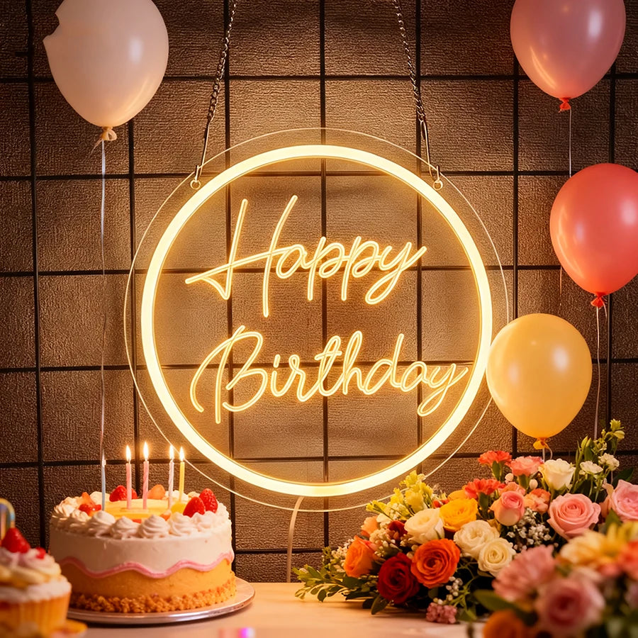 A glowing neon sign spelling Happy Birthday is displayed on a dark wall with balloons and a cake.