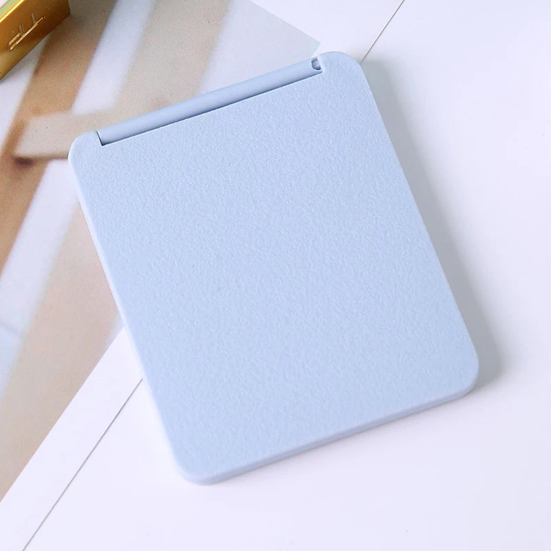 A light blue compact foldable travel makeup mirror is shown on a white surface.
