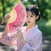A young woman in a pink silk garment holds an open pink folding hand fan decorated with red flowers.