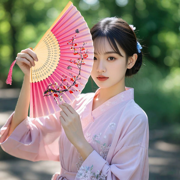 A young woman in a pink silk garment holds an open pink folding hand fan decorated with red flowers.