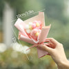 A hand holds a pink handmade crochet flower bouquet with white and pink flowers, shown against a blurred green background.