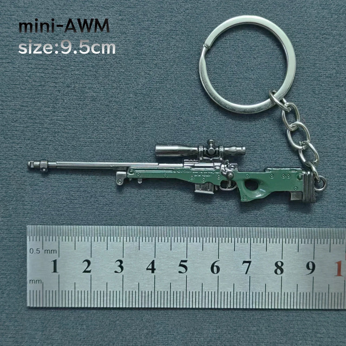 A miniature green alloy gun model keychain with a metal split ring attached, shown next to a ruler.