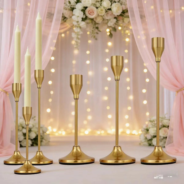 A set of elegant gold metal candle holders displayed against a soft pink draped background with floral arrangements.