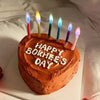 A round orange cake decorated with white lettering that reads HAPPY BOKHEE'S DAY and topped with several lit decorative candl