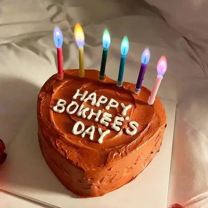 A round orange cake decorated with white lettering that reads HAPPY BOKHEE'S DAY and topped with several lit decorative candl