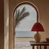 A terracotta lamp sits on a wooden table next to an arched window overlooking the sea.